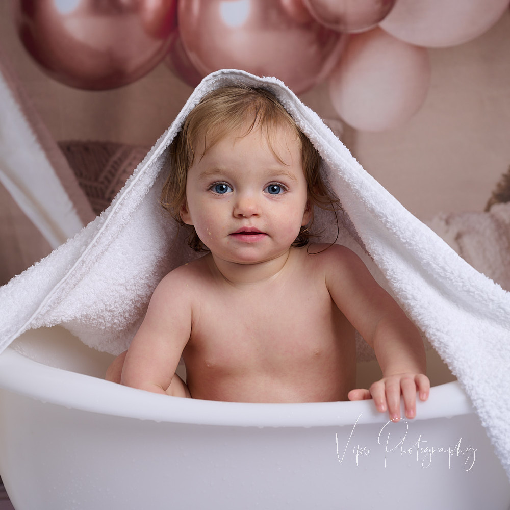 1st birthday mage of child in a bath with a pink balloon background and a towel over their head